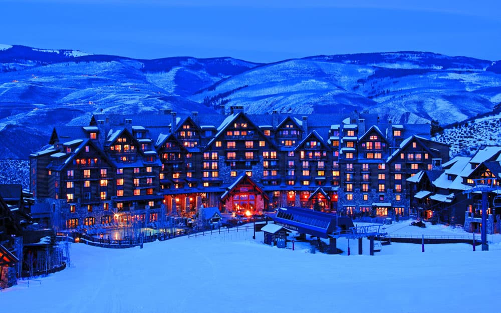 The Ritz-Carlton, Bachelor Gulch, a lodge building with many peaked roofs at dusk. Surrounded by snowy hills and covered in snow itself, the image showcases Beaver Creek in winter.