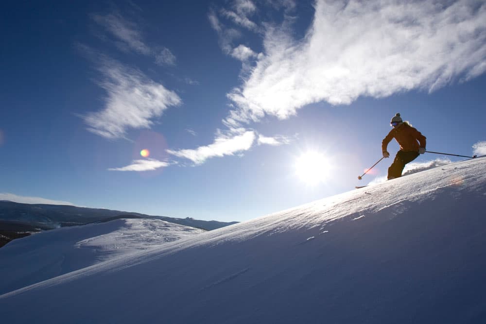 A skier skis through fresh white powder on the right with the sun in the middle of a blue sky with a few clouds.