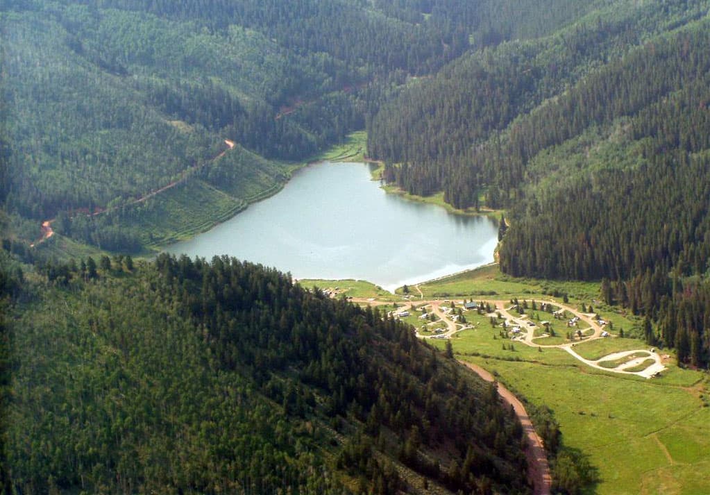 A lake is tucked into evergreen-tree covered mountains with a campground on green grass in the lower right.