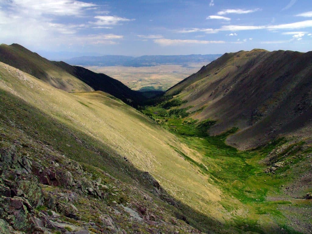 A verdant green valley lies between two barren, steep slopes near Westcliffe, Colorado. Clouds above cast shadows along parts of the valley and mountains.