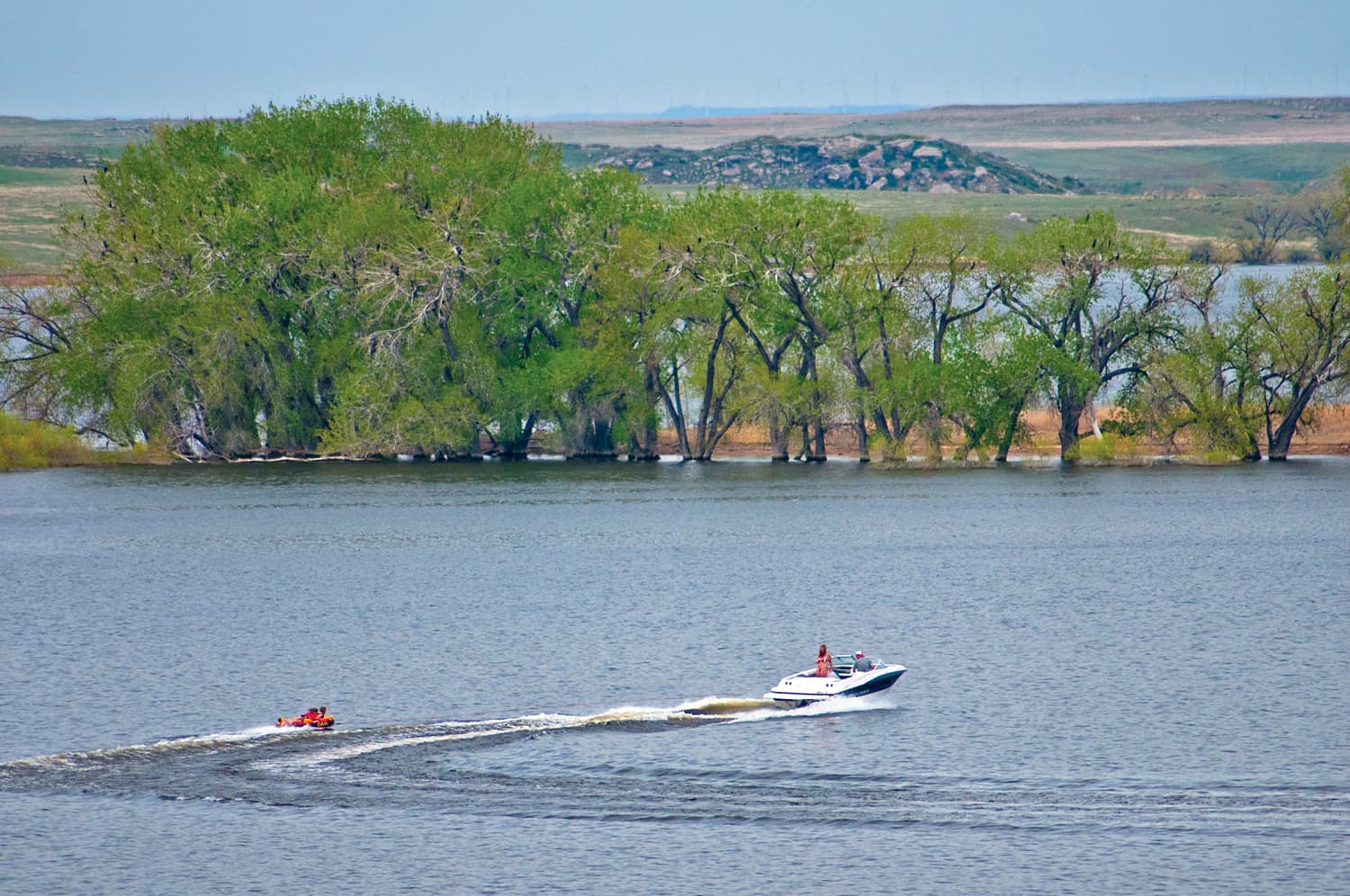 A boat pulls a tube through blue-gray water with green-leafed trees in the background.