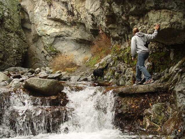 A black dog picks it way over the slippery rocks along the waterway near Zapata Falls in Colorado. A person in a gray hoodie and jeans follows behind with a camera hung over their shoulder.