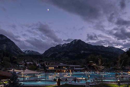 The light-blue pools of Ouray Hot Springs Pool sit under the night sky with clouds and half of the moon. There are snow-capped mountains in the distance.