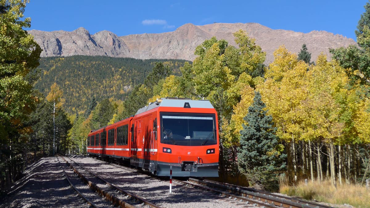 The red, squared-off Pikes Peak travels along a set of tracks next to am evergreen and golden aspen forest with Colorado mountains in the background.