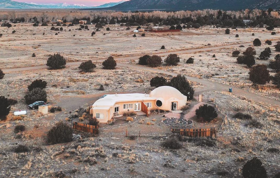 An aerial photo of the spacious dome at Crestone Bliss. The building is tan-colored and sits among flat dirt landscape speckled with dark-green shrubbery.