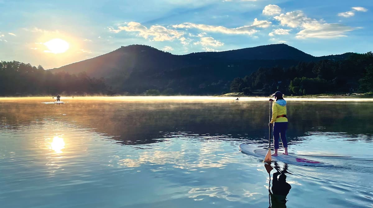 Someone is standing on a stand-up paddleboard on Evergreen Lake at sunset. The waters ripple gently behind them as their paddle goes through the water. In the background are towering mountains under a party cloudy sky.