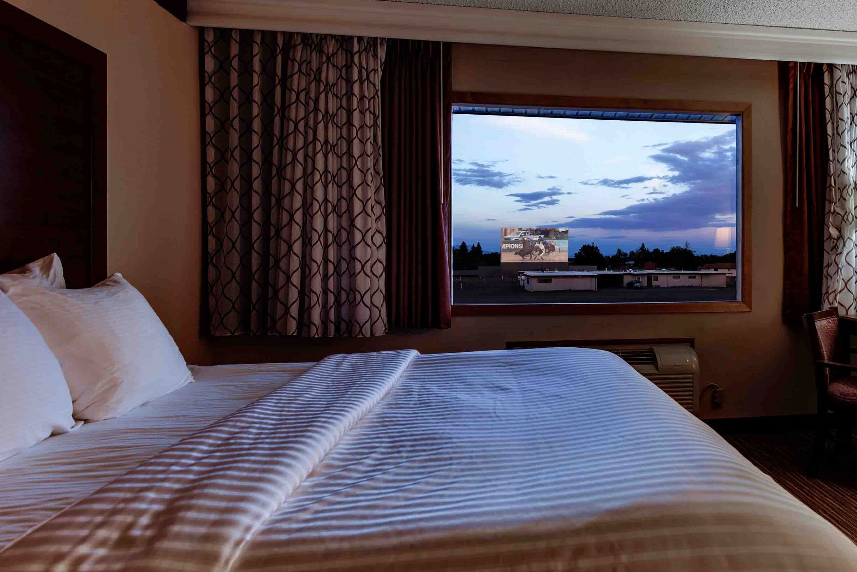 The curtains on a hotel window are pulled open to reveal a drive-in movie screen, visible from the comfort of a cozy, white-blanketed bed at Best Western Movie Manor in Monte Vista Colorado.