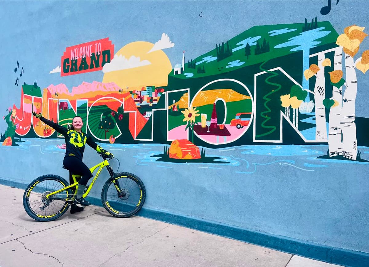 Someone on a bike poses next to a mural in Downtown Grand Junction, which says "WELCOME TO GRAND JUNCTION." There is a cartoon sun, rock formations, birds, aspen trees, flowers and lakes that decorate the mural.