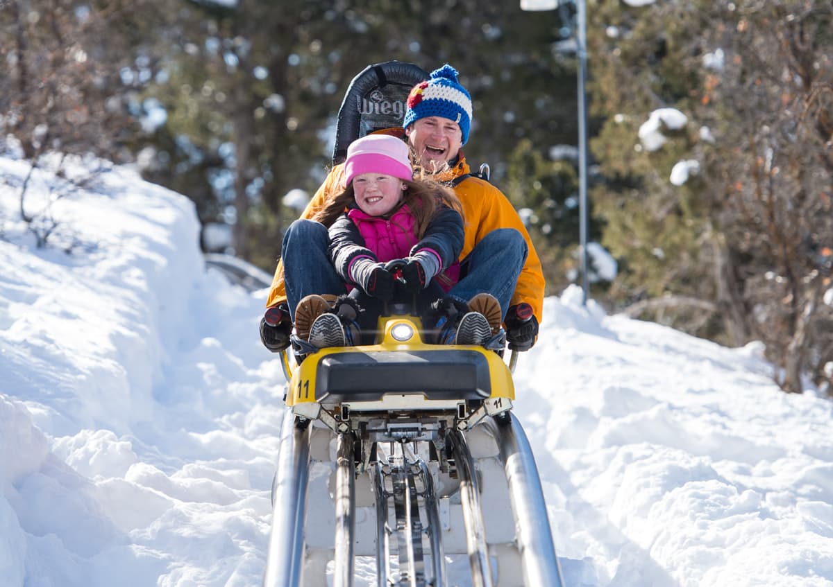 A parent with a child bundled up in winter gear and riding in front of them on an alpine coaster which is cutting through the snow.