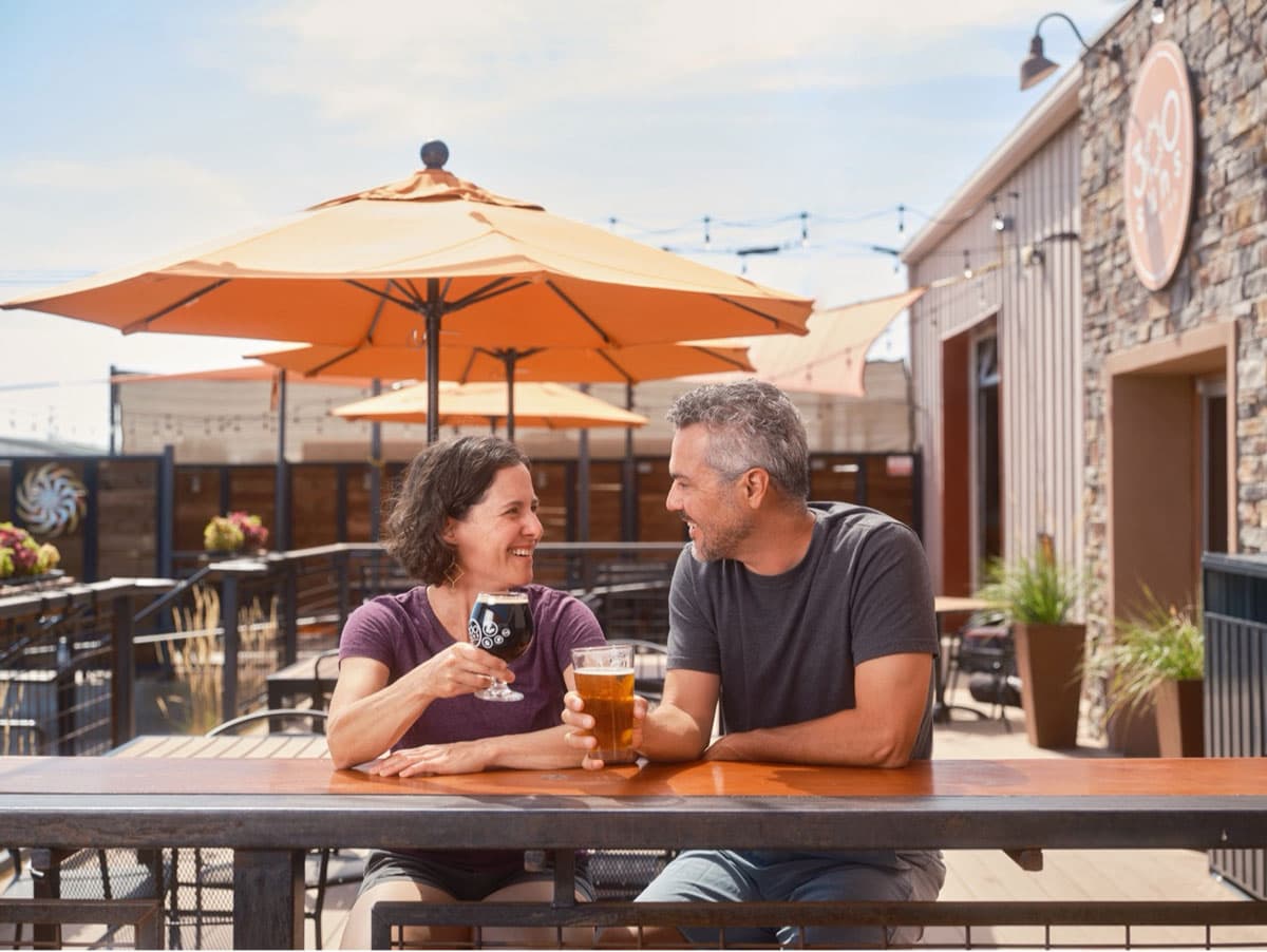 Smiling couple sharing drinks at a brewery patio with outdoor seating.