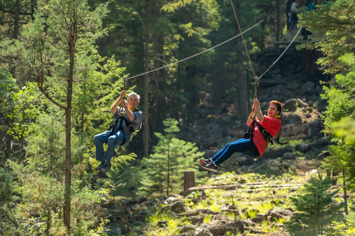 A man and woman ziplining side-by-side in a forested area in Colorado.