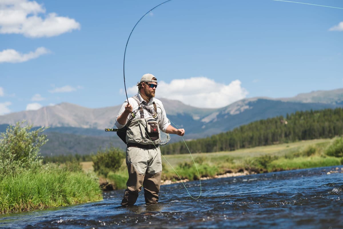 A man fly-fishing in the middle of a river with snowy mountains in the background
