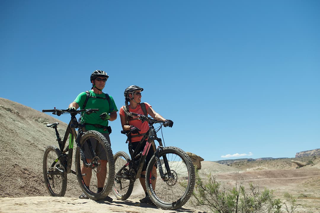Two mountain bikers in helmets standing on a dirt landscape in Grand Junction, holding onto their bikes and looking out into the distance. The sky above them is a dusty blue.