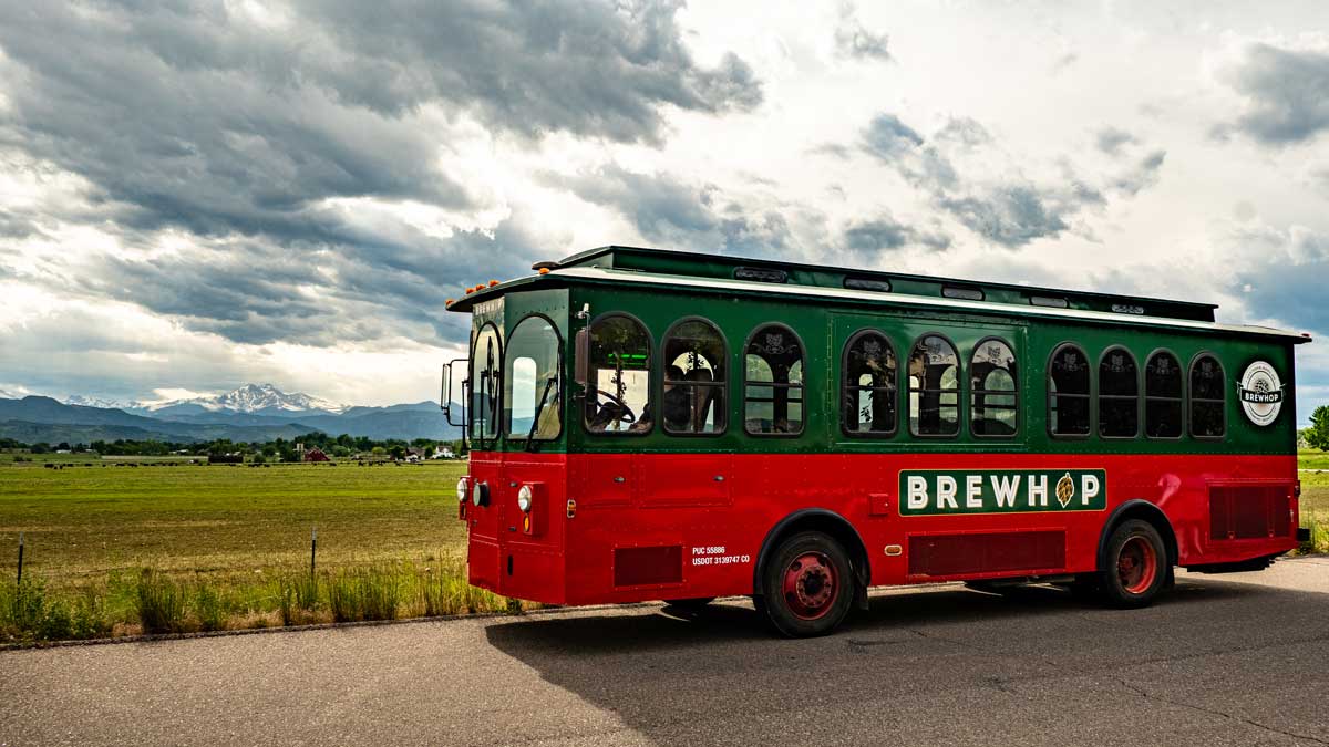 The green and red BrewHop Trolley makes its way down a grey stretch of road with green farm land in the background and in the hazy distance a snow-capped mountain sits.