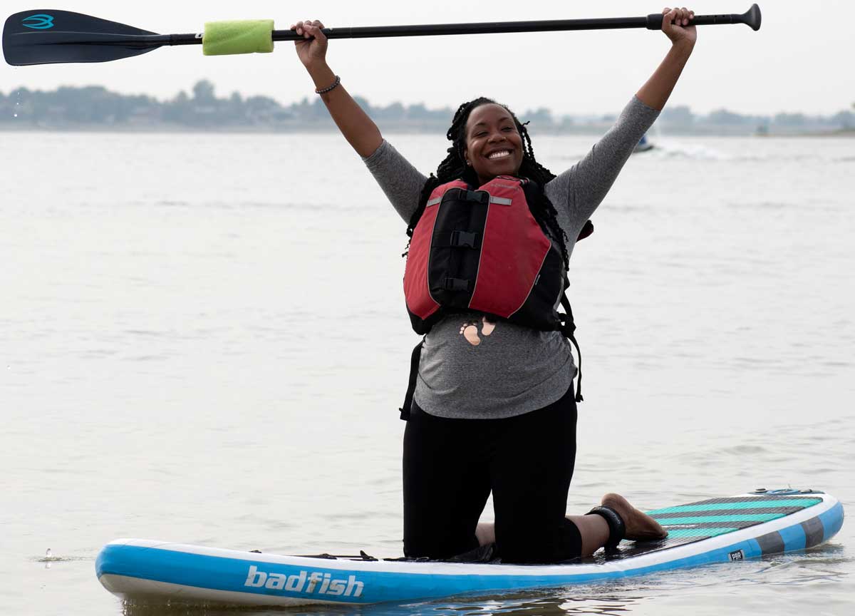 A water adventurer kneels on a standup paddleboard near Loveland, Colorado. They raise their paddle above their head and smile.