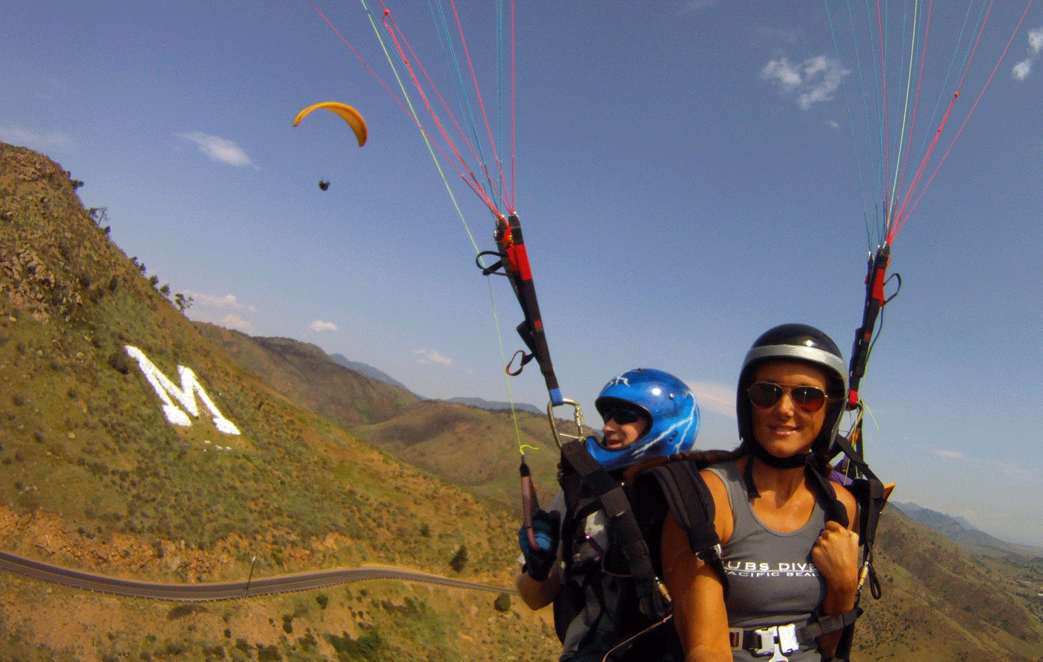 We see two helmeted paragliders swinging gracefully from their parasail. Beyind them, we see an "M" outlined on a hill behind them (it stands for the school of Mines that's in Golden); another paraglider under an orange parasail can also be seen floating behind them