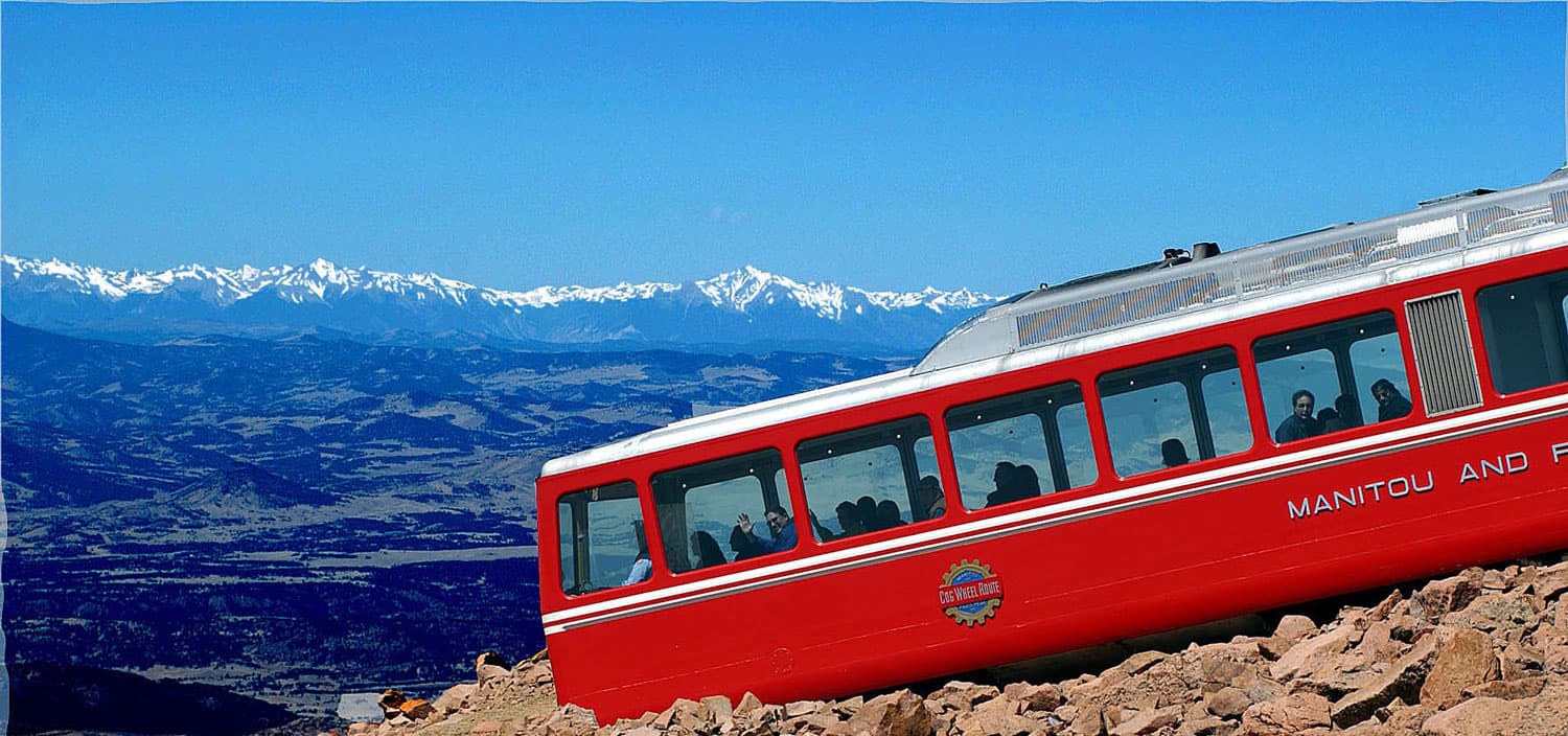 A bright red passenger compartment on a new train summits the rocky mountainside of America's Mountain in Colorado. Snow-capped peaks appear in the distance.