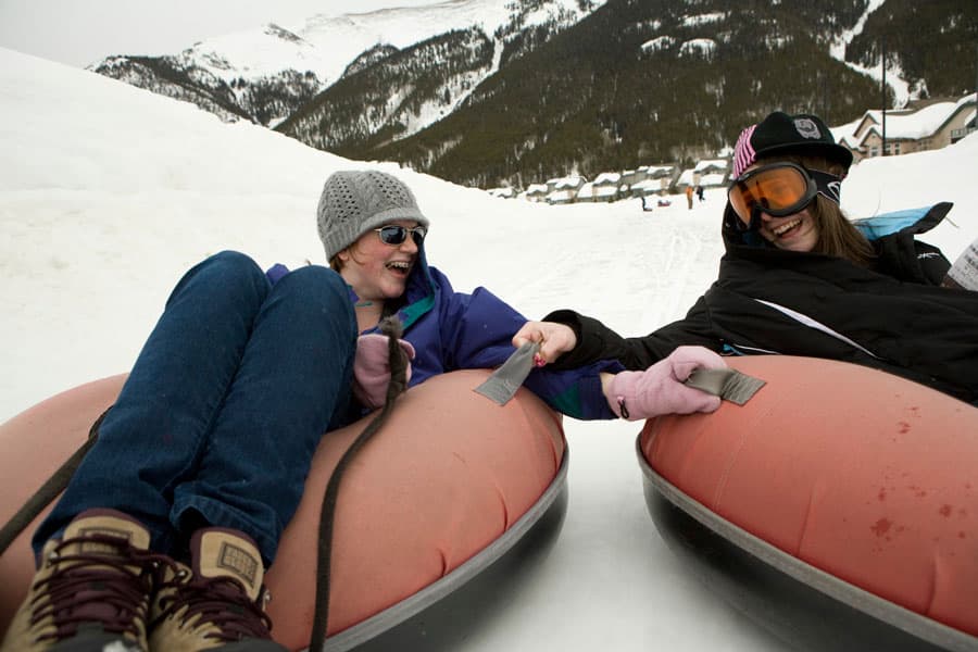 Two children hold hands while snow tubing down a mountain