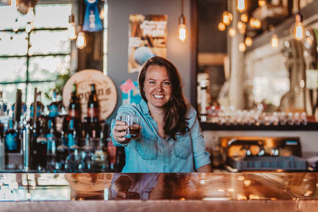 A bartender in a blue shirt holds up a glass of liquid at the copper bar at Abbott & Wallace Distilling in Longmont