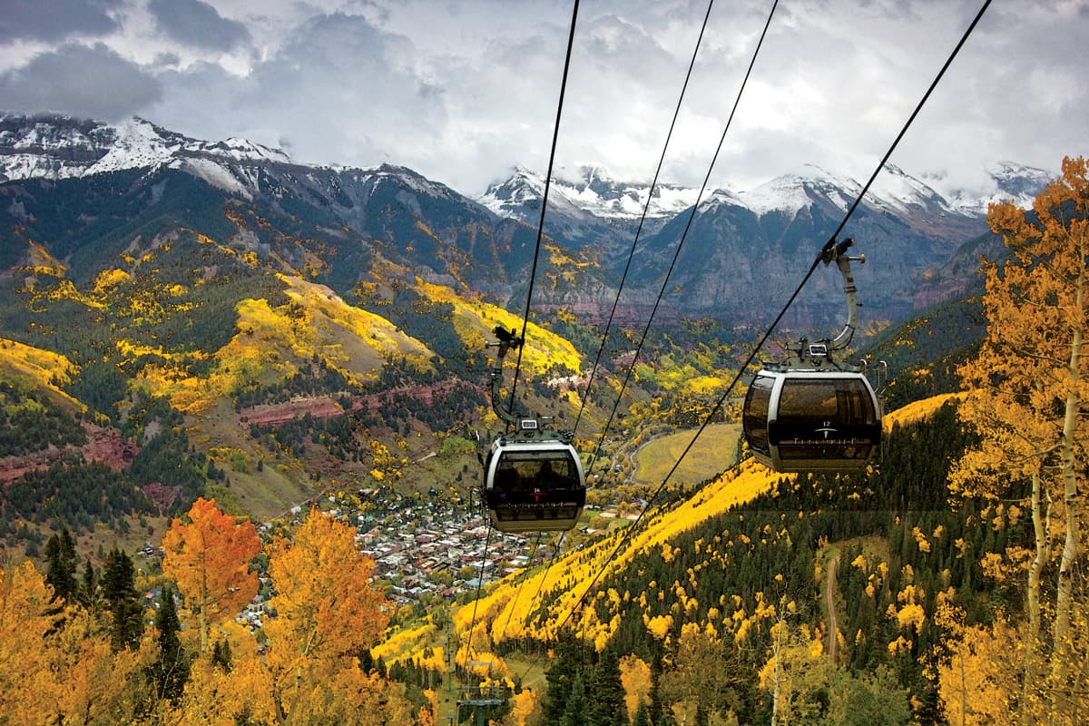 Two silver gondolas with large tinted windows travel up a mountainside on a metal cable. Snow-capped mountain shrouded in clouds and vibrant fall colors near Telluride, Colorado, are seen in the background.