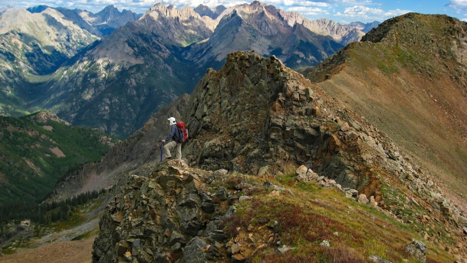 Someone in climbing gear and a helmet stands perched on top of the summit of Twilight Peaks in Weminuche Wilderness near Durango, Colorado. All around them are stunning, jagged peaks stretching as far as the eye can see under a mostly sunny sky.