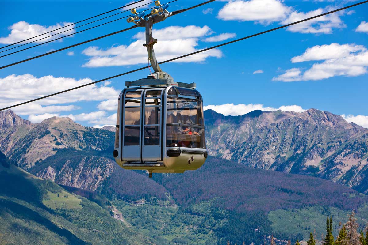 A gondola with large windows travels along a wire cable near Vail, Colorado. The nearby scenery is breathtaking with blue skies and rugged, lush, green mountains.