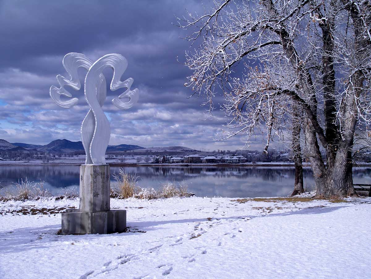 A sculpture sitting on the snow-covered ground surrounding McIntosh Lake. There's a tree covered in snow on the right and in the cloud-covered sky there's mountain peaks in the distance.