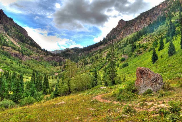 A winding path leads through a green-grass valley with evergreen trees. On both sides Rocky Mountain peaks soar toward a cloud-covered blue sky.