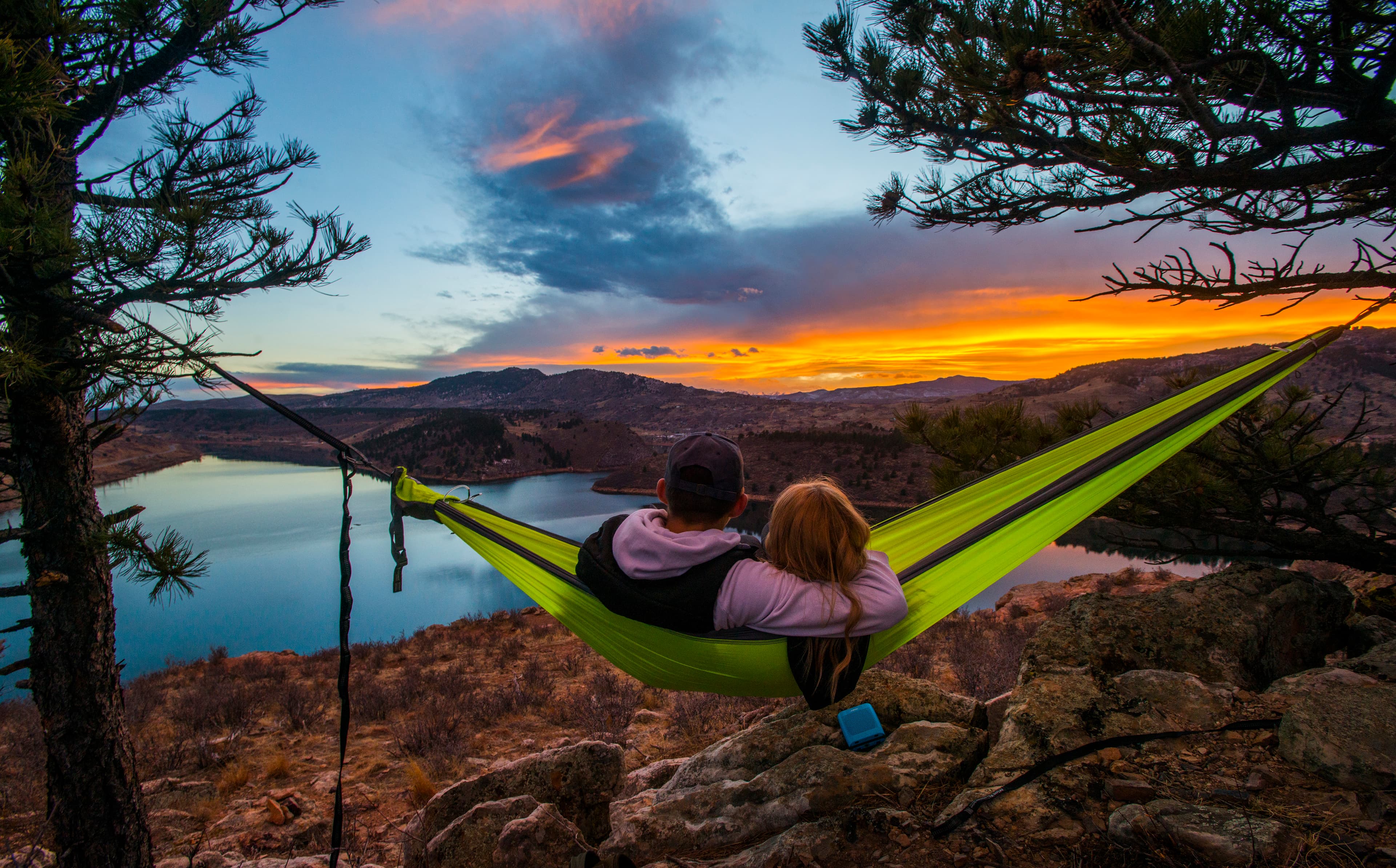 A couple in a lime-green hammock, that's strung up between two pine trees above a rocky outcropping look out onto Horsetooth Reservoir at sunset. In the distance a mountain meets an orange sky that's tinged with blue.