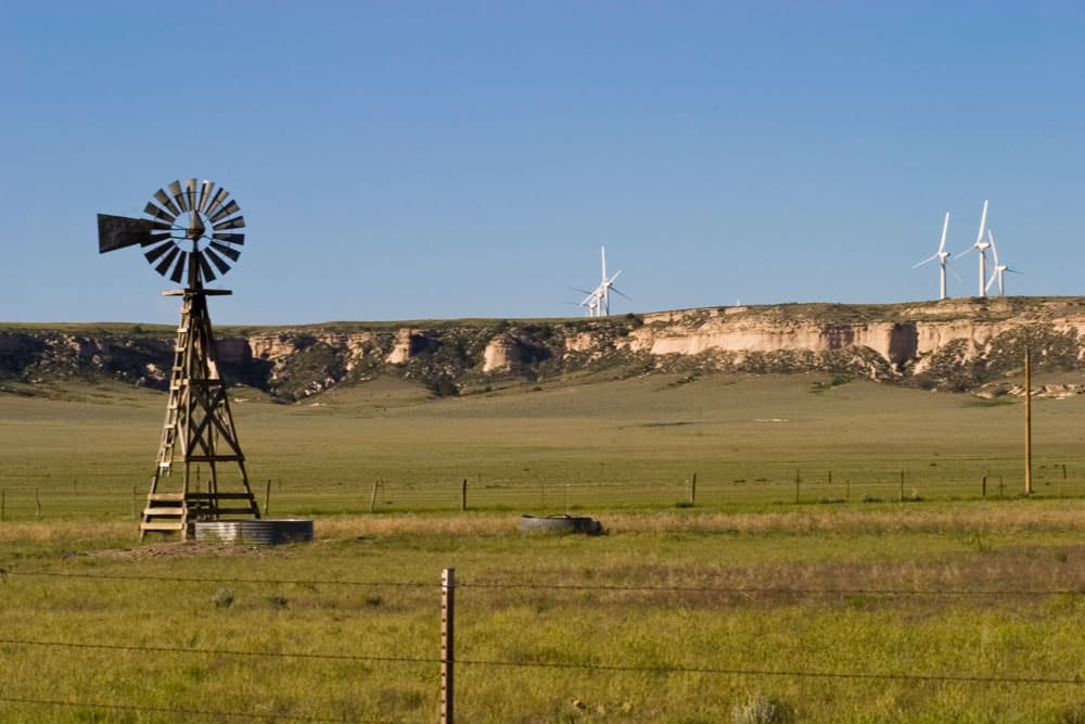 An old-fashioned windmill stands in a field at the Pawnee National Grassland in Colorado. On the other side of a rocky ridge, several modern, sleek air turbines spin.