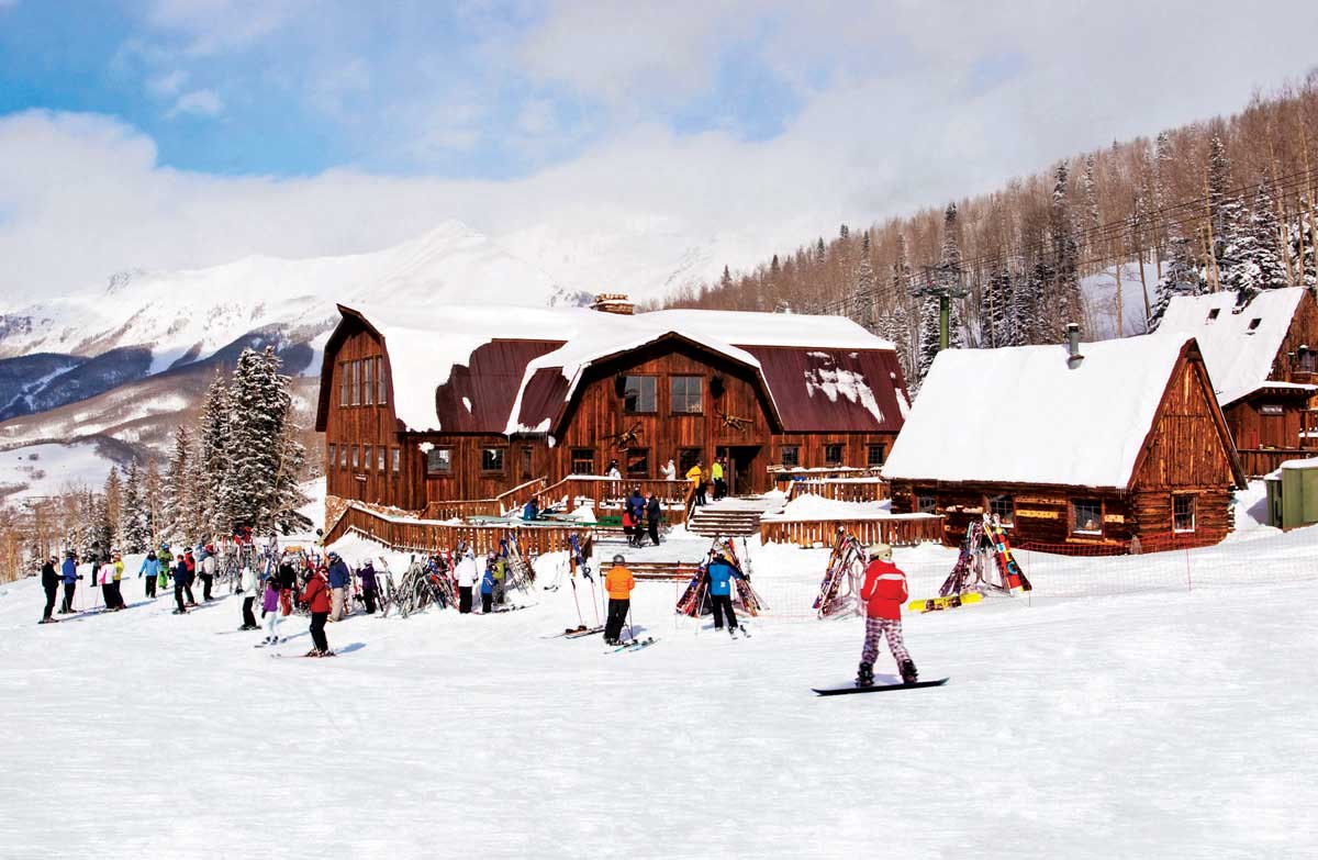 Skiers and snowboarders gather in the snow around the outside of a large wooden lodge at Telluride Ski Resort.