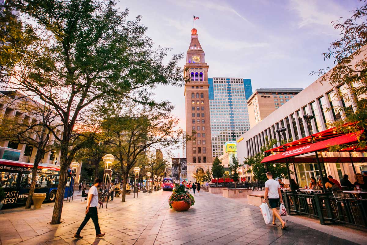 A panoramic photo of 16th Street in Denver, lined with lively outdoor seating at restaurants and tall buildings of downtown. The sky is the clear-blue of summer.