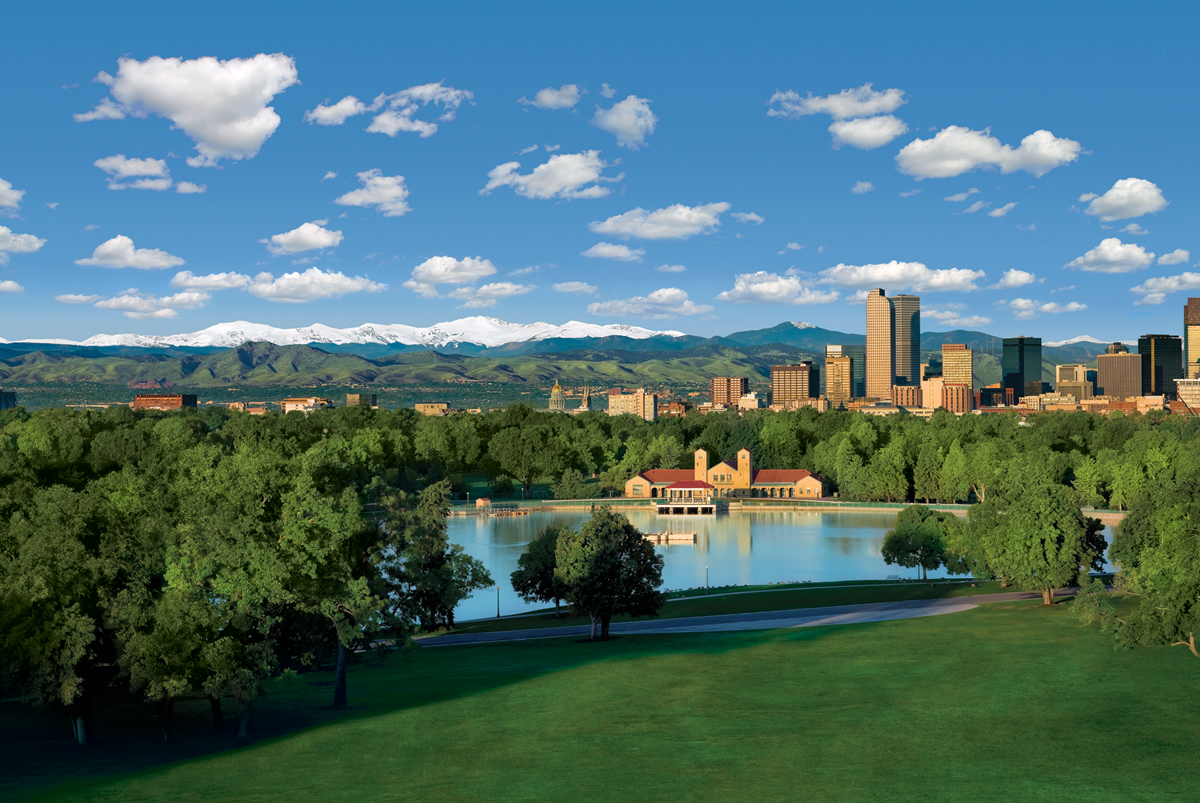 A photo looking over City Park, Ferril Lake, the City Park Pavillion and the downtown Denver skyline on a summer day. The Rockies are visible on the horizon underneath cloud-spotted blue skies.