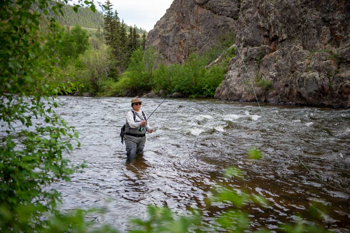 Through the green trees, a woman is fly-fishing on the Taylor River in Almont. There are rock structures and green trees behind her.