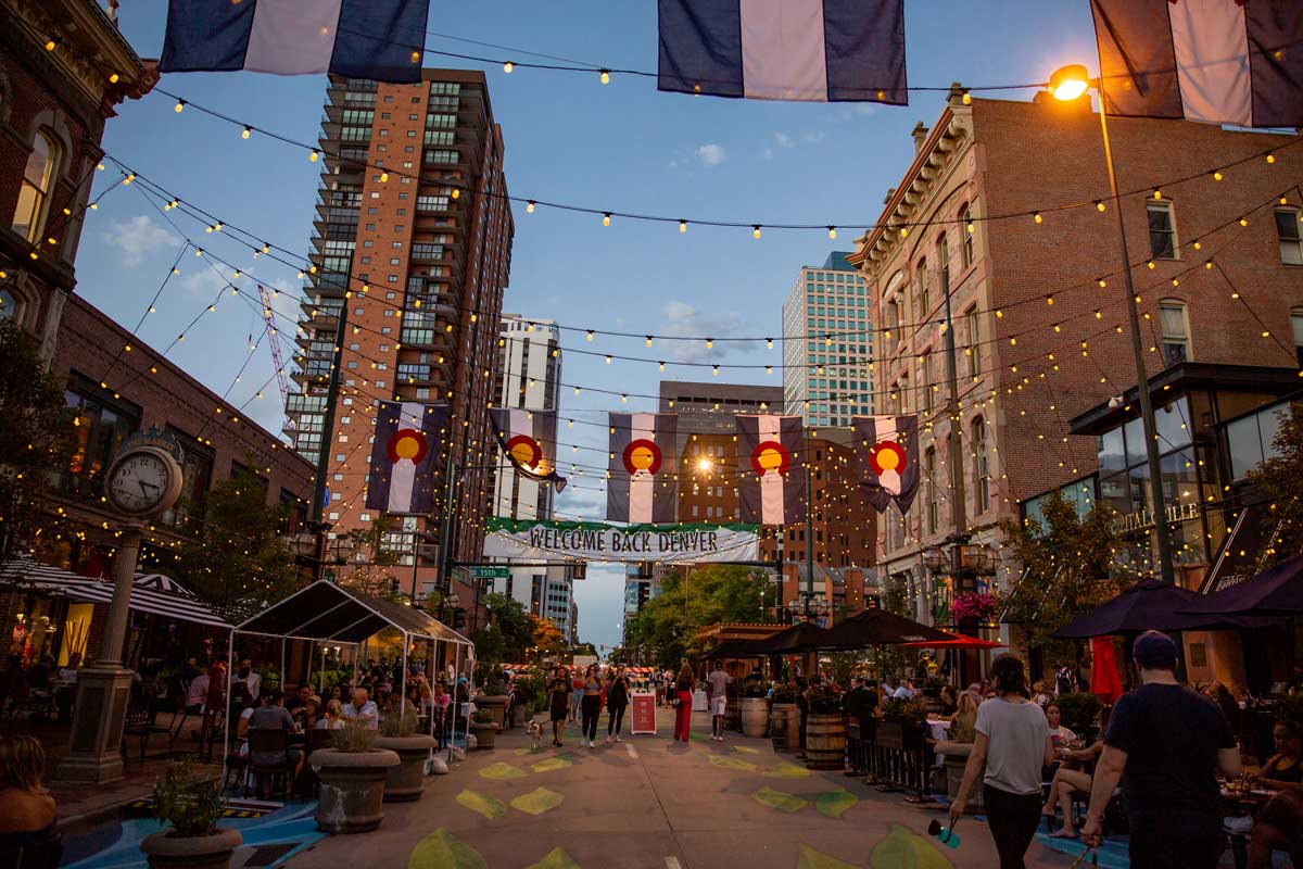 A busy walkway street on Larimer Square in downtown Denver, with buildings surrounding the walkway strung with fairy lights. People are walking and chatting between the restaurants during a clear summer evening.