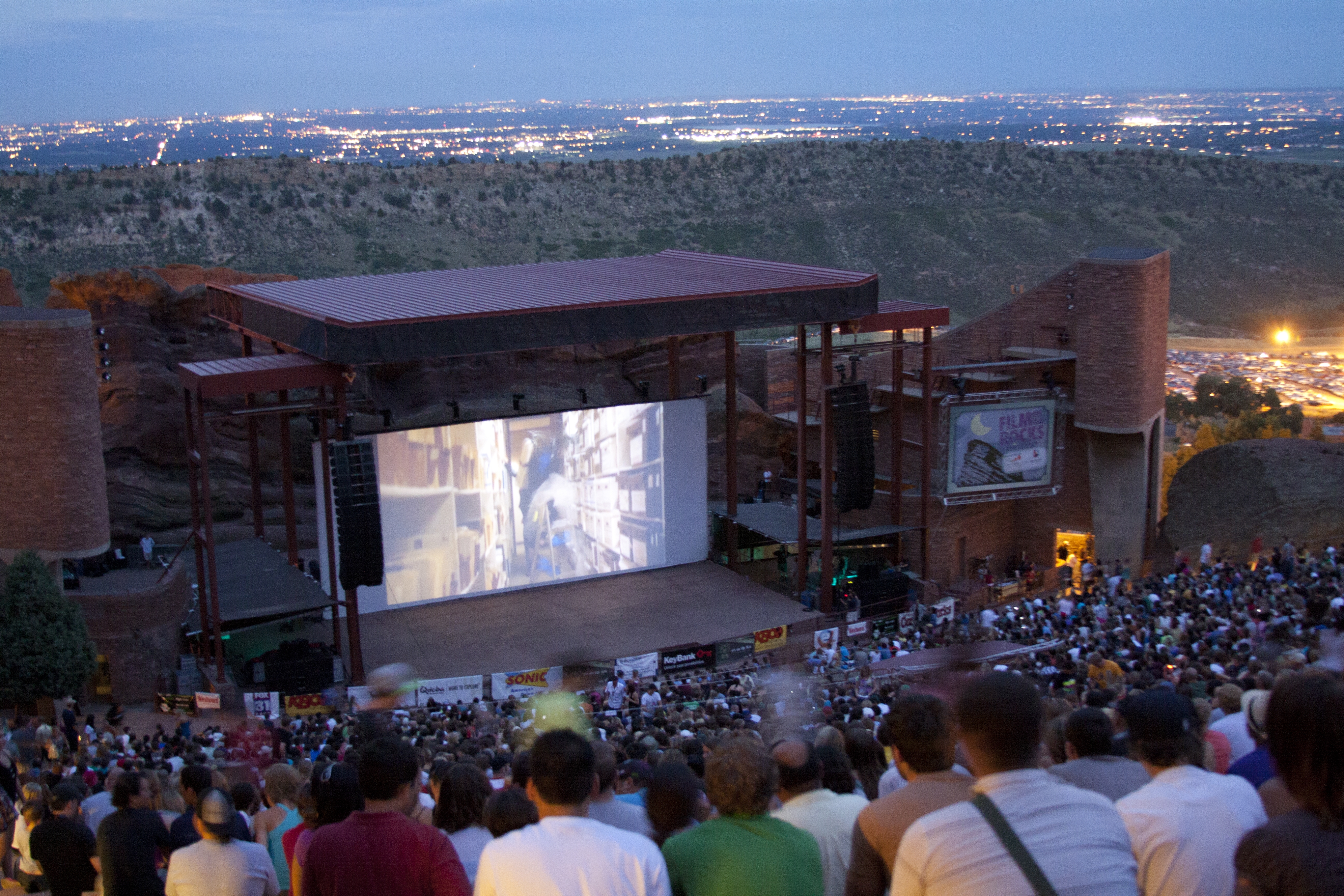 A crowd watches a film on an outdoor stage surrounded by mountains