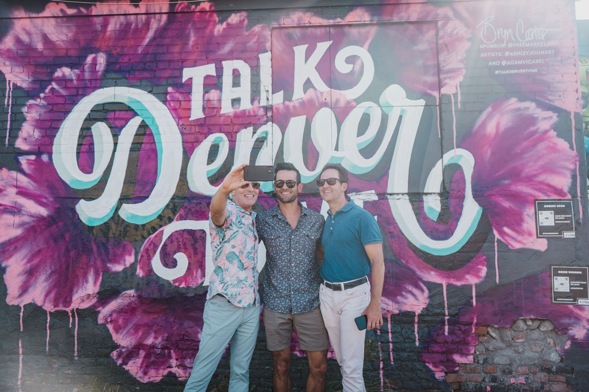 Three people pose before a floral Talk Denver to Me mural in Denver's RiNo neighborhood in Colorado.