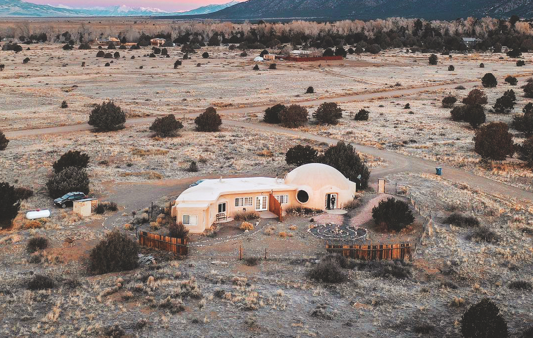 An aerial photo of the spacious dome at Crestone Bliss. The building is tan-colored and sits among flat dirt landscape speckled with dark-green shrubbery.