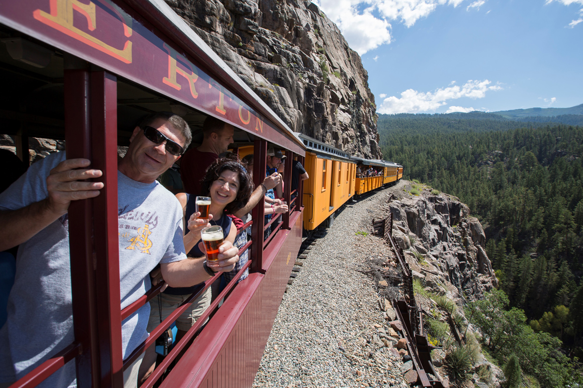 Two people holding small glasses of ruby-red beer pose in an open-air passenger car on a Durango Train as the train moves along tracks on a narrow, gray-rock cliffside.