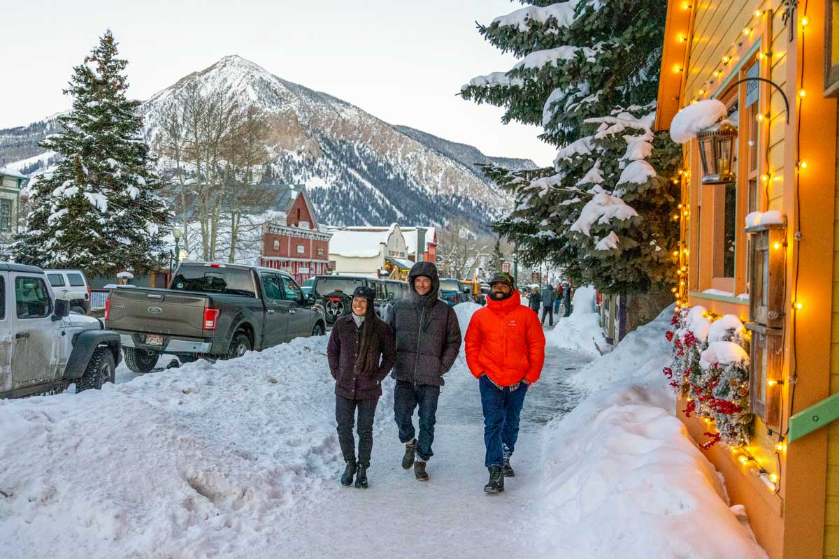 Three people bundled in winter coats walk down a snoy sidewalk on Gunnison's main street. We can see a snow-covered hill behind them and piles of shoveled snow on each side, as well as lit up storefronts