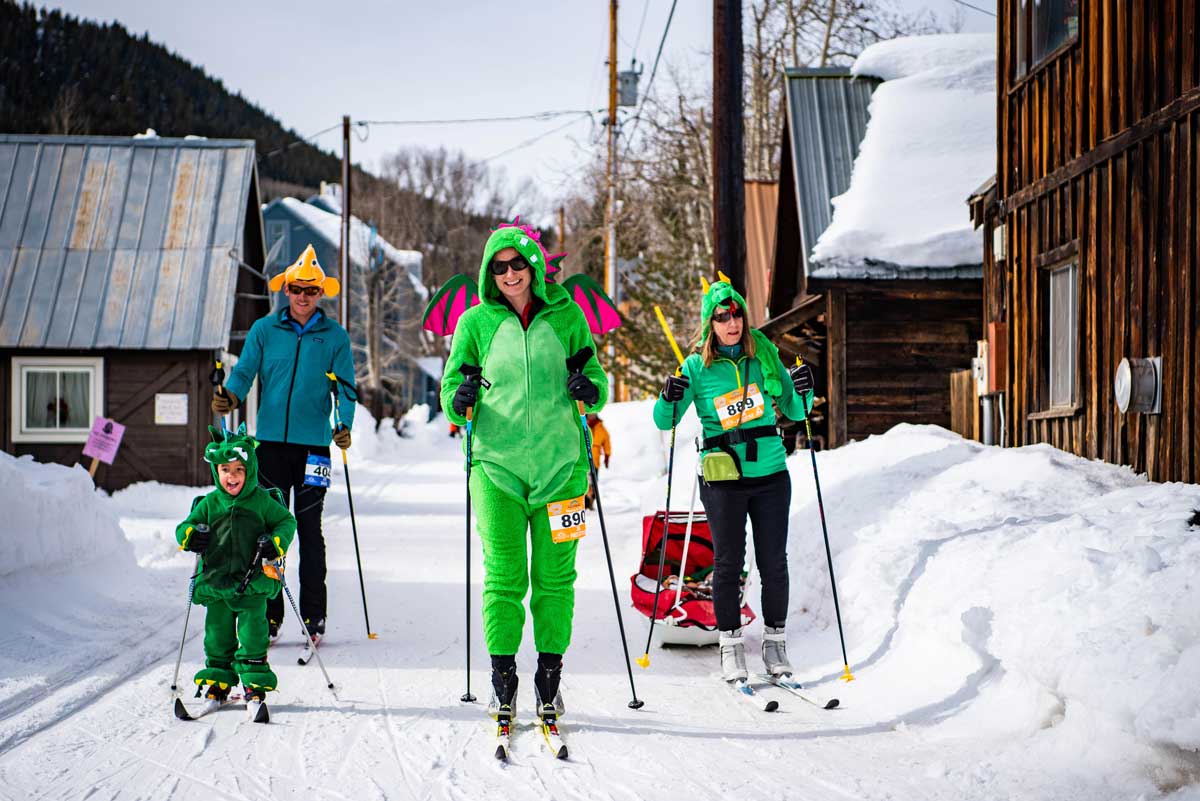 Four Nordic skiers dressed up green onsies use poles to scoot down a snowy road between houses with snow-covered rooftops
