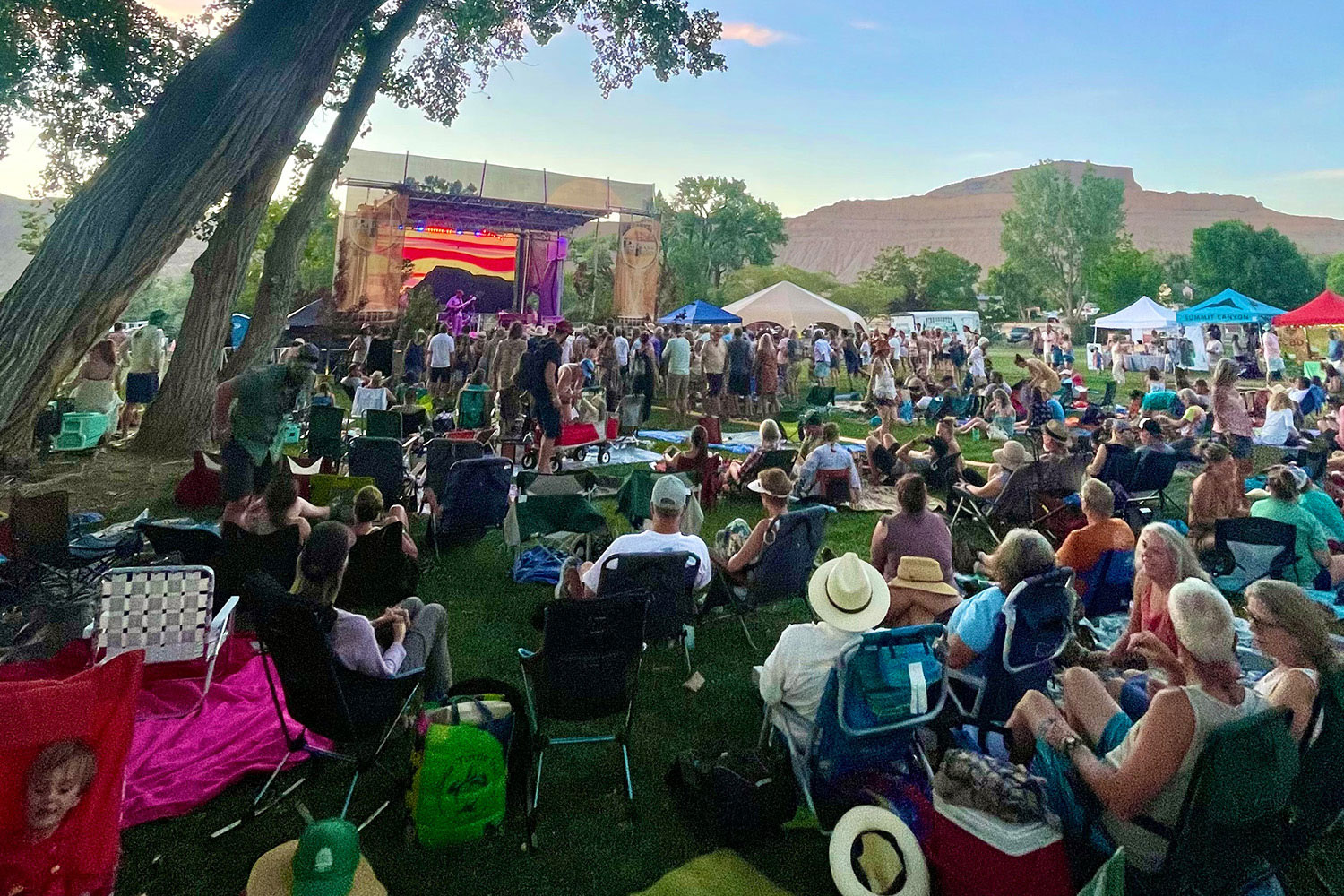 A crowd of people sitting on a lawn face a stage at the Palisade Bluegrass Festival