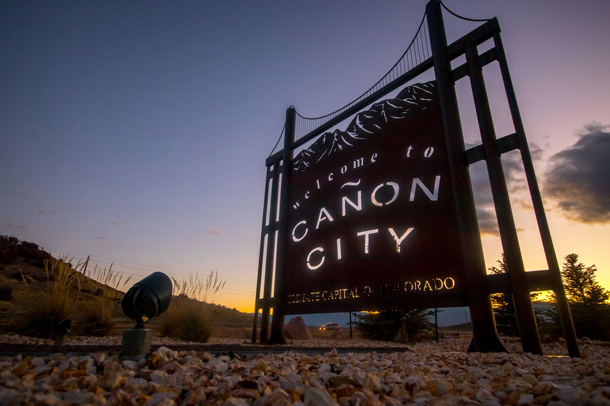 A tall metal sign with a replica of the Royal Gorge that says "Welcome to Cañon City." In the background is a deep purple and yellow sunset with minimal clouds.