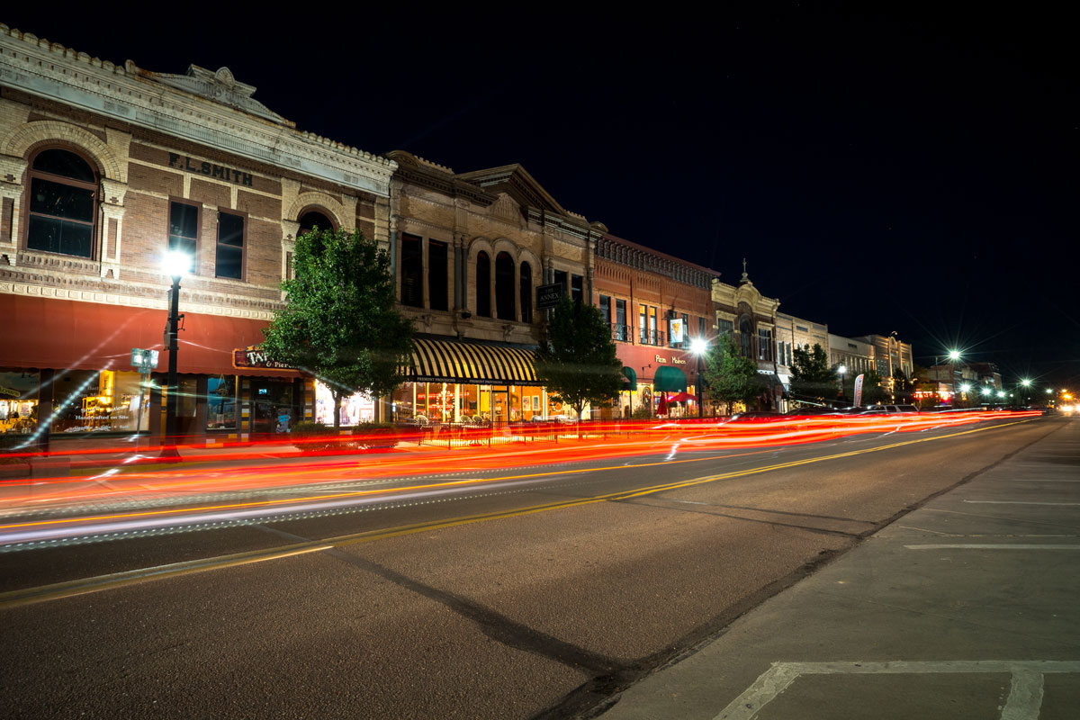 A strip of storefronts in downtown Cañon City at night. Red lights from cars in motion make a few steady streaks above the road.