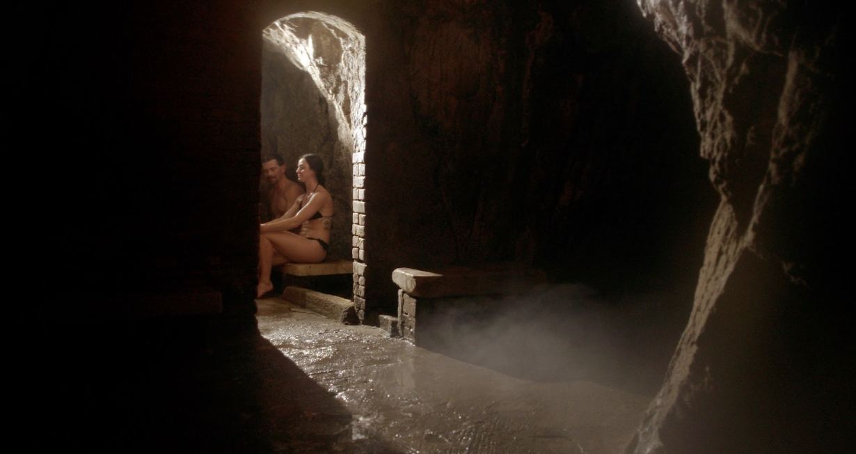 A couple wearing swimsuits sits on a wooden bench in a dimly lit cave and breathe in the steamy vapors at a spa in Colorado.