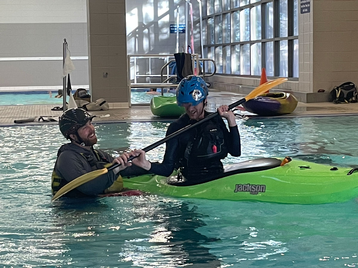 A student sits in a green kayak inside an indoor pool. They are wearing protective gear and are holding a double-sided paddle. Their instructor is in the water next to them. They are both smiling. In the background is another pool illuminated by tall floor-to-ceiling windows letting sunlight beam in.