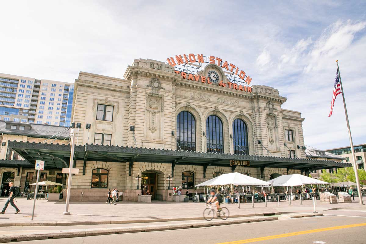 Under softly lit skies, Denver's Union Station rises majestically, while a patio bar and pedestrians walk in front of it
