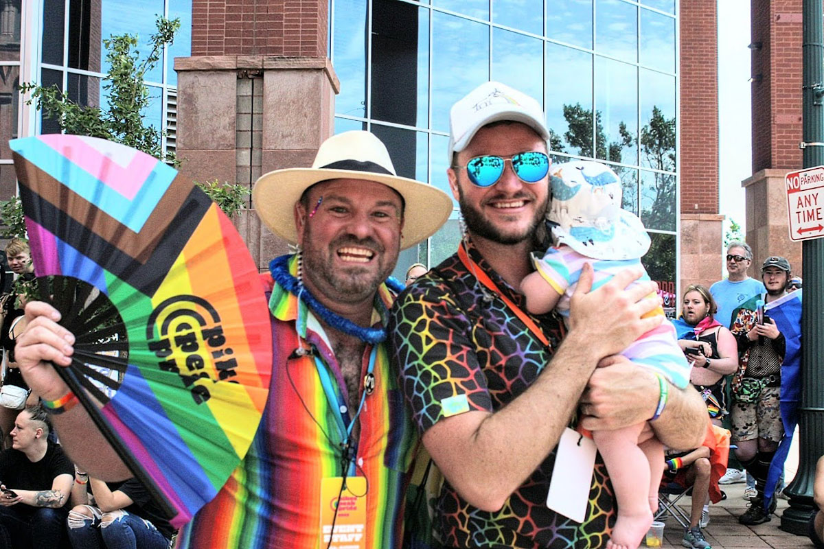 Two people pose together while smiling widely and wearing rainbow-colored clothes. One of them is holding a pride folding fan and the other holds a baby in a bucket hat.