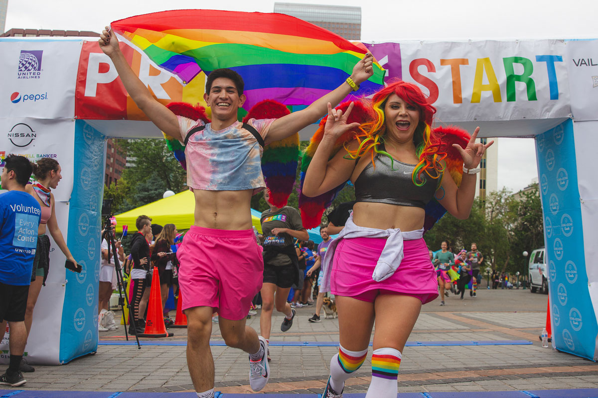 Two joyous people wearing rainbow wings on their backs run across the finish line of the Denver PrideFest 5K race. One of them holds a pride flag tapestry behind them.