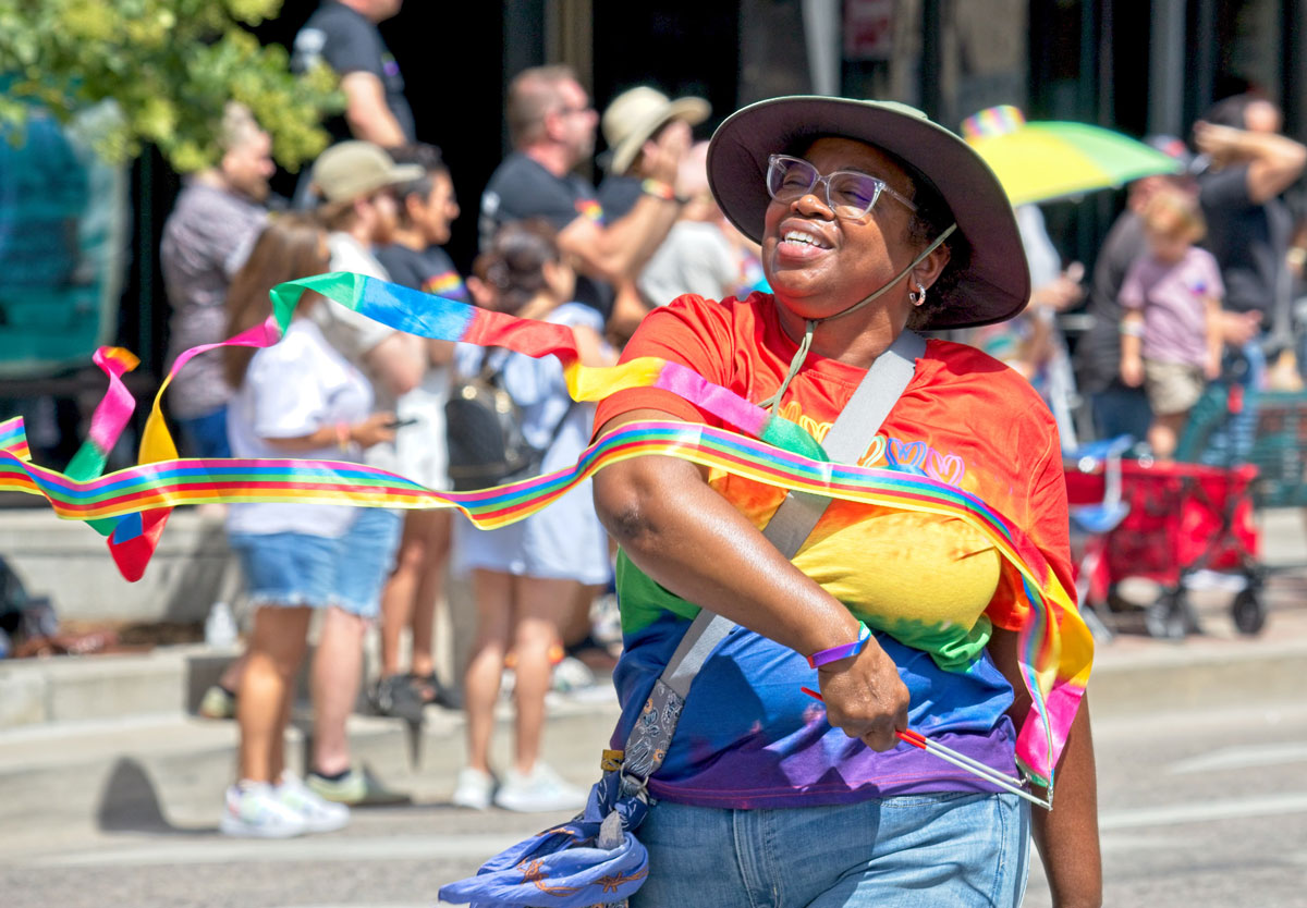 A person in glasses, a bucket hat, a rainbow t-shirt and jeans waves long rainbow ribbons on sticks in front of them as they walk in a pride parade in Colorado Springs.